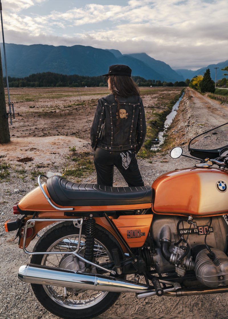 Person standing next to an orange motorcycle with mountains in the background wearing Soul Sister since 1969