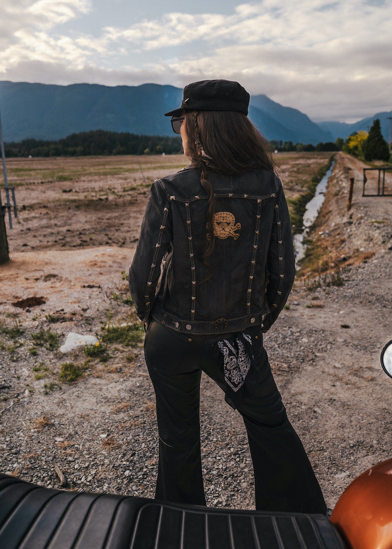 Person wearing a black jacket with skull design and cap, standing on a dirt road with mountains in the background wearing clothing designed and made by Soul Sister since 1969..