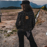 Person wearing a black jacket with skull design and cap, standing on a dirt road with mountains in the background wearing clothing designed and made by Soul Sister since 1969..