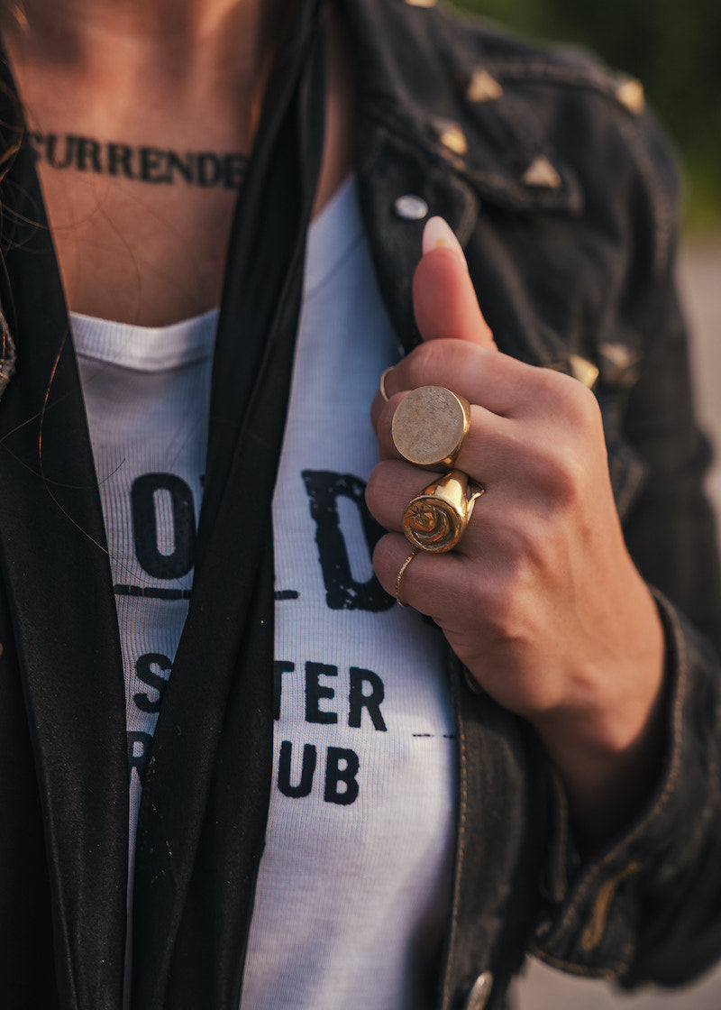 Person wearing a black jacket with gold buttons, white shirt with text, and multiple rings on a blurred background wearing clothing designed and made by Soul Sister since 1969..