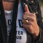 Person wearing a black jacket with gold buttons, white shirt with text, and multiple rings on a blurred background wearing clothing designed and made by Soul Sister since 1969..