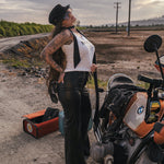 Woman in a white tank top and black pants standing next to a motorcycle on a dirt road wearing clothing designed and made by Soul Sister since 1969.