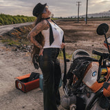 Woman in a white tank top and black pants standing next to a motorcycle on a dirt road wearing clothing designed and made by Soul Sister since 1969.