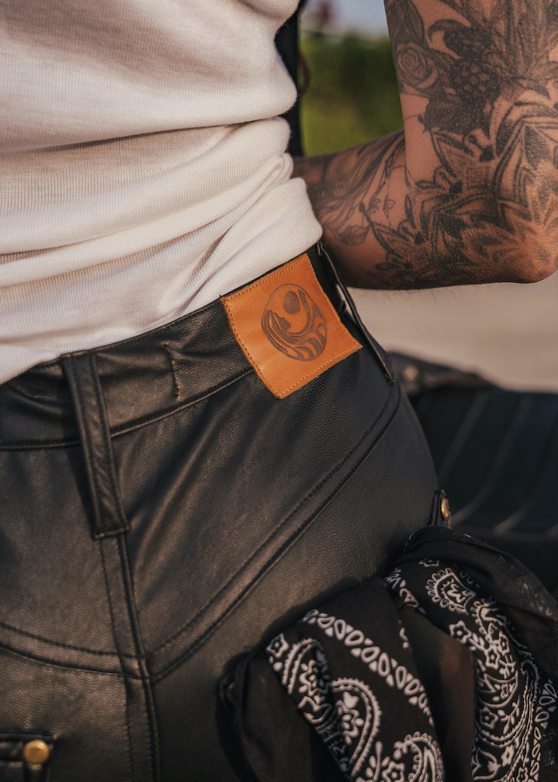 Person wearing black leather pants with a brand patch, tattooed arm visible wearing clothing designed and made by Soul Sister since 1969.