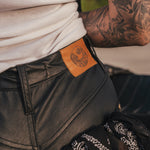 Person wearing black leather pants with a brand patch, tattooed arm visible wearing clothing designed and made by Soul Sister since 1969.