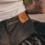 Person wearing black leather pants with a brand patch, tattooed arm visible wearing clothing designed and made by Soul Sister since 1969.