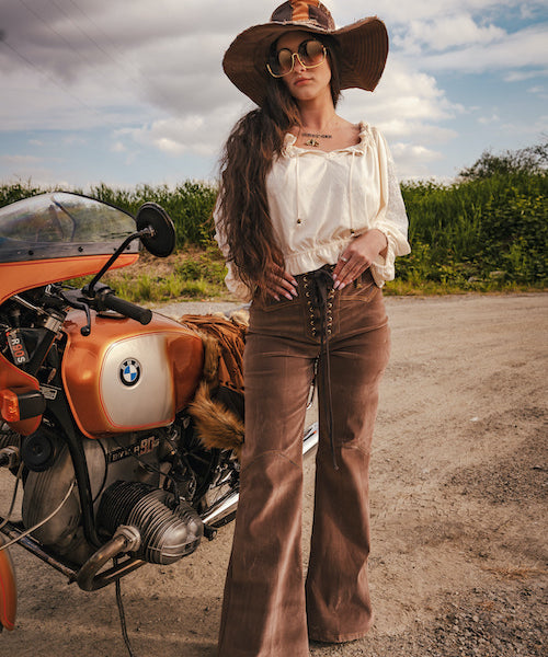 Woman in vintage attire standing next to a BMW motorcycle on a road with a scenic background wearing Soul Sister since 1969