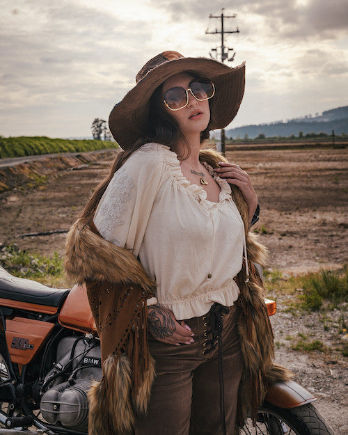 Person in rustic attire with a fur vest and wide-brimmed hat standing next to a motorcycle in an open field. wearing Soul Sister since 1969