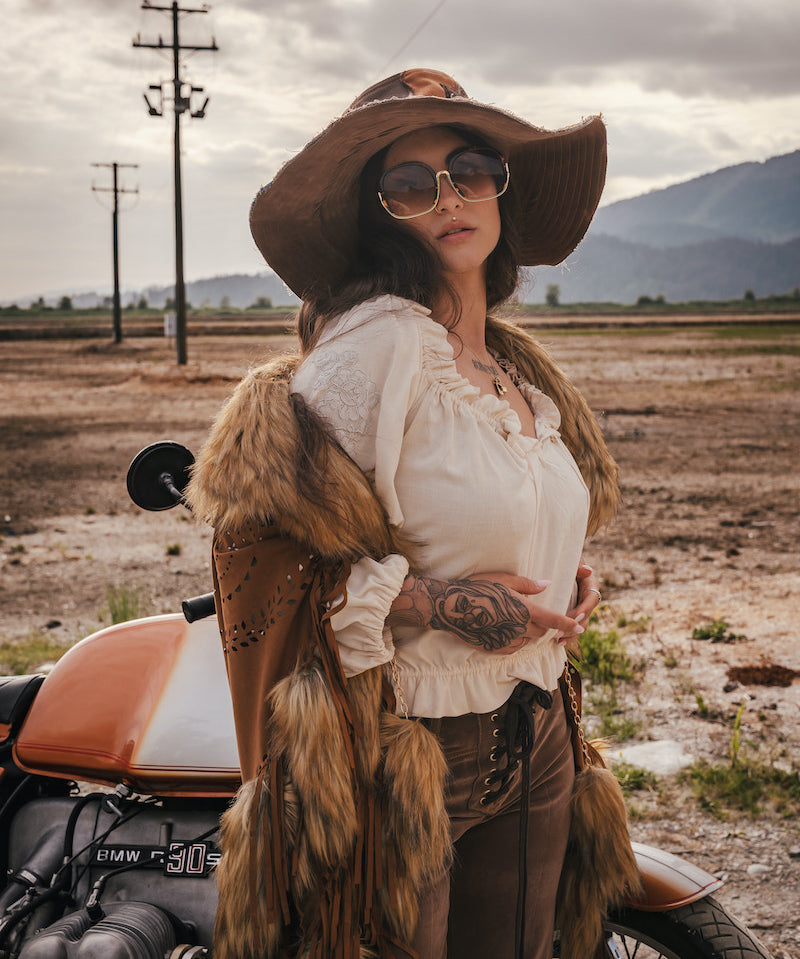 Woman in rustic attire with fur vest and wide-brimmed hat standing next to a motorcycle in an open field wearing Soul Sister since 1969.