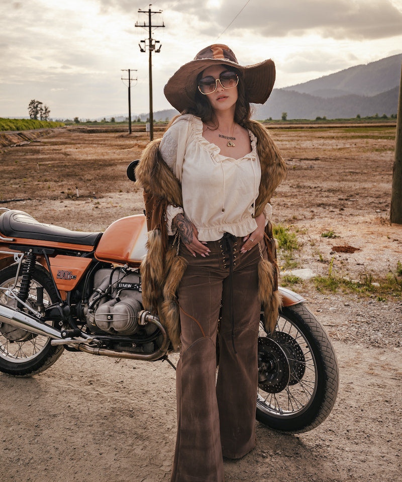 Person in vintage attire standing next to a motorcycle in an open field with mountains in the background wearing Soul Sister since 1969