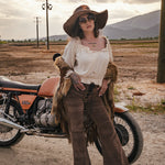 Person in vintage-style clothing standing next to a motorcycle in an open field with mountains in the background. wearing Soul Sister since 1969