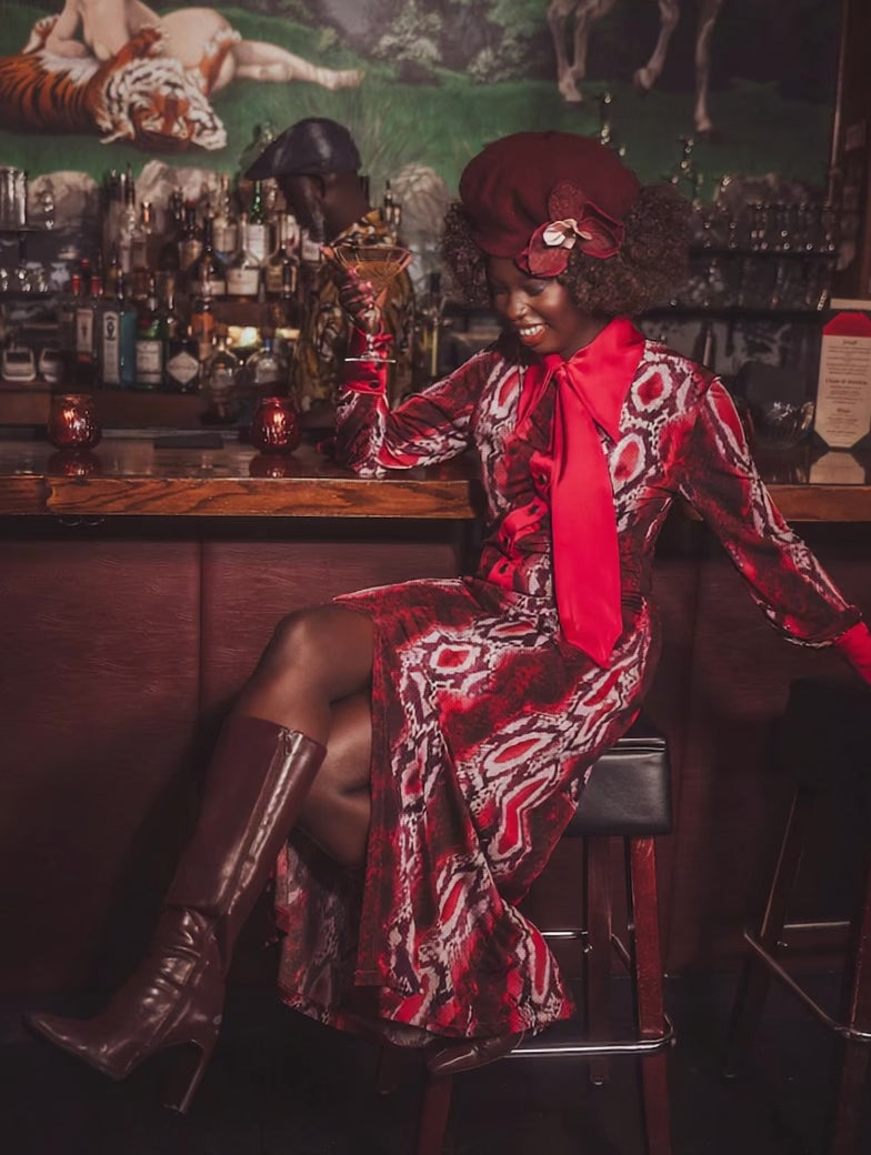 A woman sits at the bar with a martini in her hand in a speakeasy setting wearing a cool 70s inspired retro dress in snake print