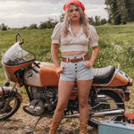 Woman standing next to a vintage motorcycle in a field wearing Soul Sister since 1969.