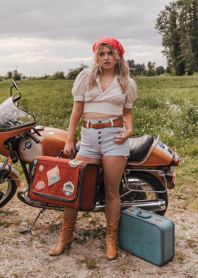 Woman standing next to a motorcycle with a suitcase in a field wearing Soul Sister since 1969.